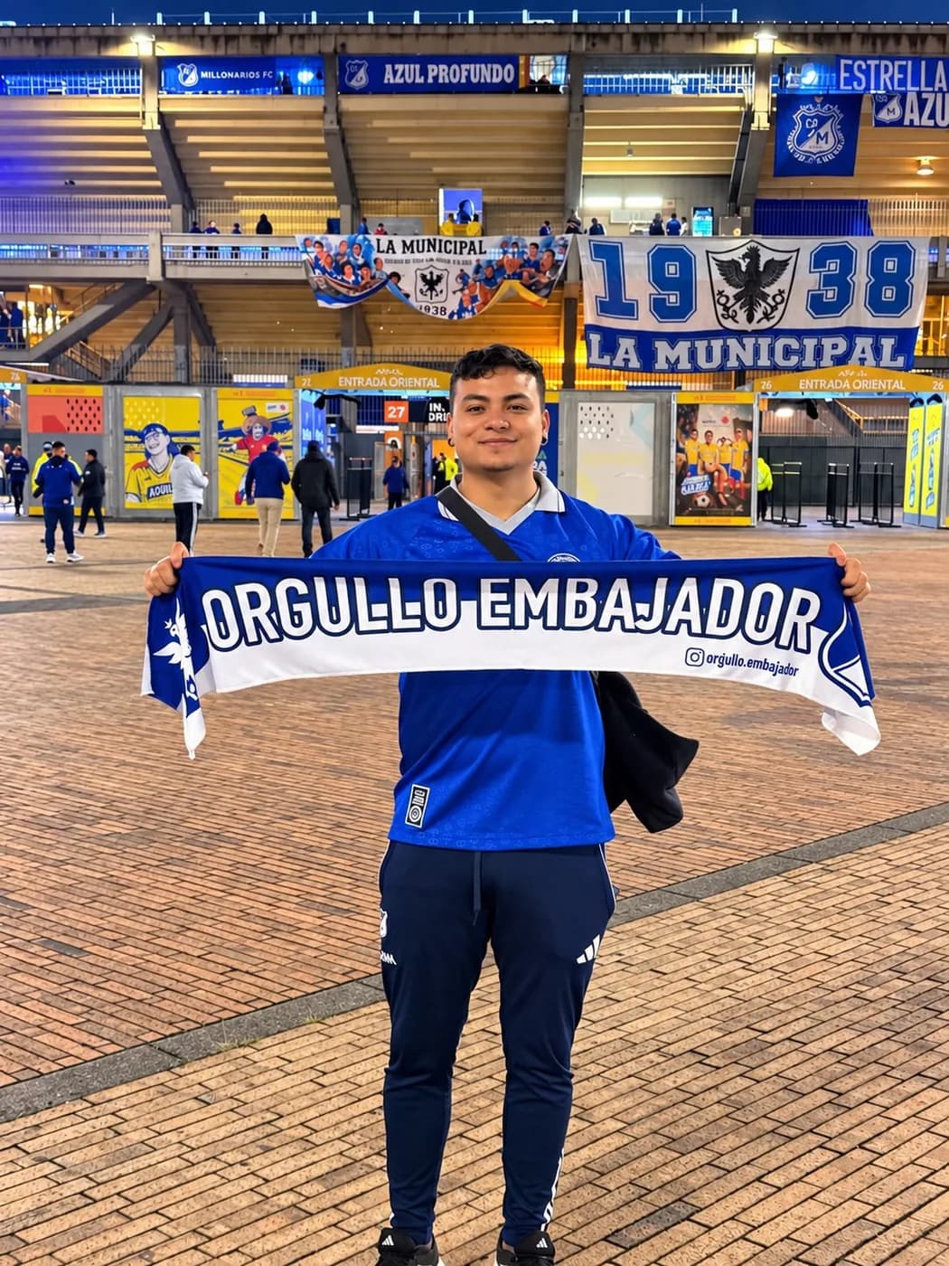 Hincha con la camiseta azul sosteniendo la bufanda de Orgullo Embajador frente a la entrada oriental de El Campín, de noche