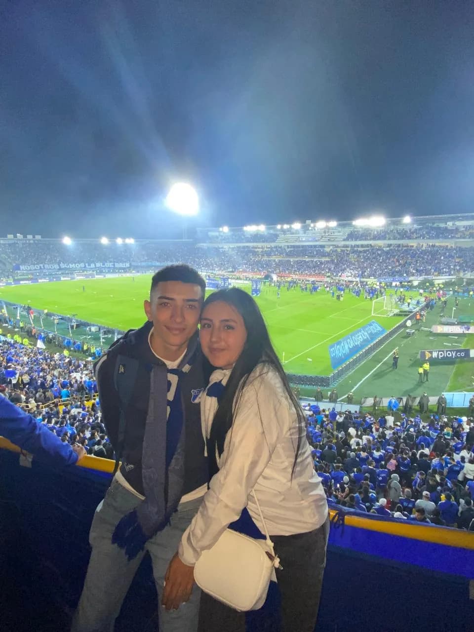 Pareja abrazada en la tribuna alta de El Campín, con la cancha iluminada de fondo