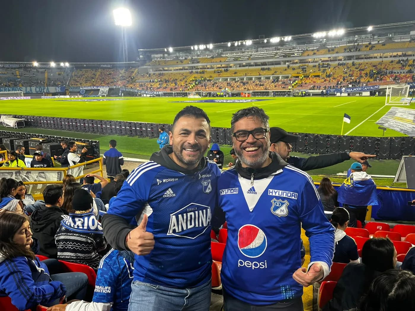 Dos amigos con camisetas azules de Millonarios junto a la cancha de El Campín, de noche