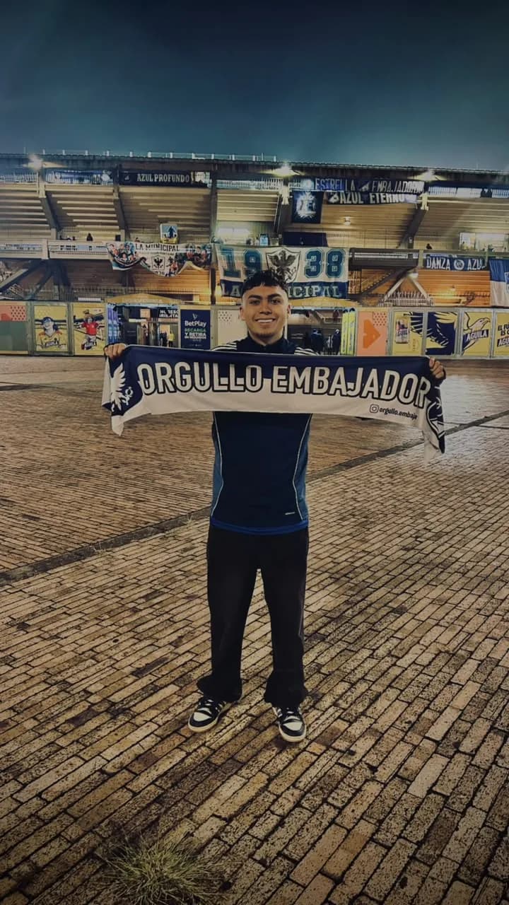 Hincha sonriendo con la bufanda de Orgullo Embajador frente a la tribuna de El Campín, de noche