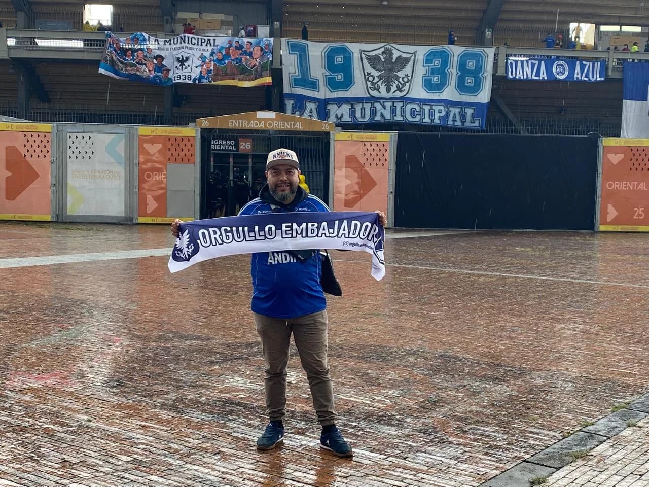 Hincha con gorra y la bufanda de Orgullo Embajador en la plaza de El Campín bajo la lluvia