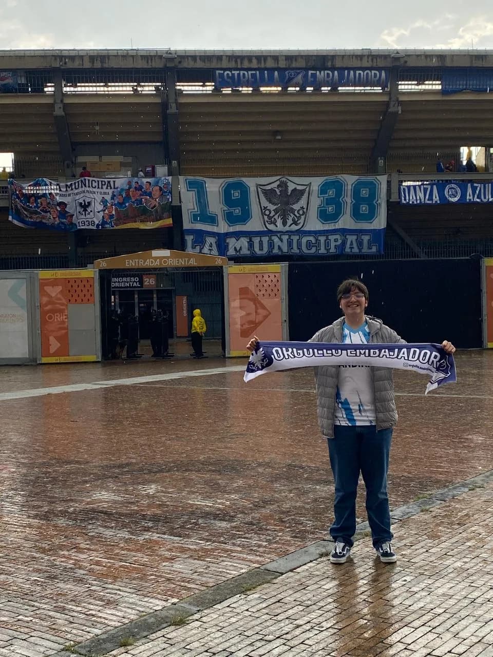 Hincha con la bufanda de Orgullo Embajador frente al trapo de 1938 en El Campín, bajo la lluvia